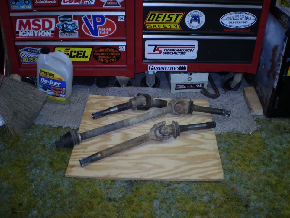 Three front axle shafts with U-joints on plywood in front of a red toolbox.