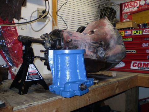 1978 Ford Bronco - A red and black automatic transmission is supported by a black jack stand and a blue part on a wooden workbench. Various tools and storage…