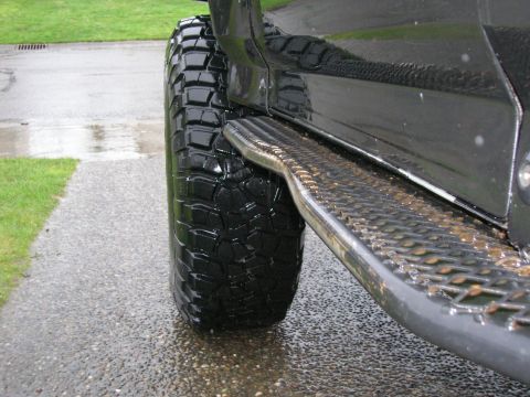 1996 Ford Bronco - A close-up side view of a black Ford Bronco's large, aggressive tread tire and diamond plate step bar. The vehicle is parked on a wet, grav…