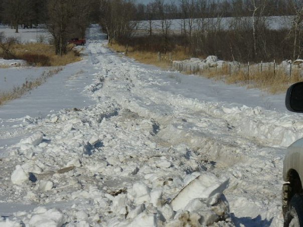 Snowy road with deep ruts and a red truck in the distance. Part of a white vehicle is visible on the right