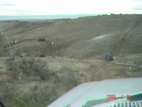 1983 Ford Bronco - Two off-road vehicles navigate a dirt track in a desolate, hilly terrain. Image has a watermark.