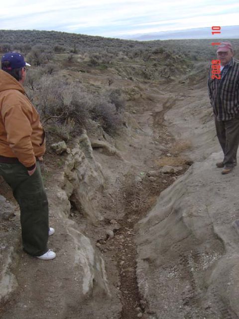 1983 Ford Bronco - Two men stand on the edge of a rocky ravine, surveying the dry creek bed below under an overcast sky.
