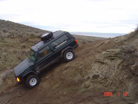 1983 Ford Bronco - Jeep Cherokee XJ navigates a steep, muddy trail with a spare tire on its roof.