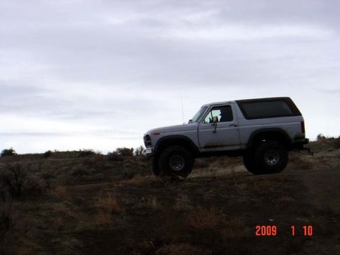 1983 Ford Bronco - A silver Ford Bronco is parked on a dirt incline with sparse vegetation and a cloudy sky overhead.