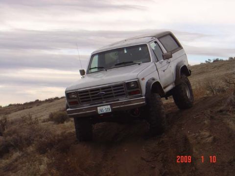 1983 Ford Bronco - A white 1980s Ford Bronco navigates a steep, muddy incline under an overcast sky.
