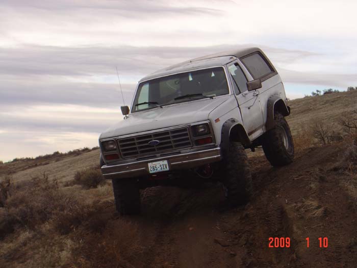 White 1980s Ford Bronco with aftermarket wheels and tires driving on a dirt hill