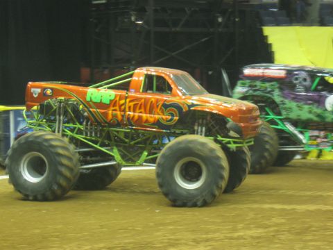 1994 Ford Bronco - Two monster trucks are parked on a dirt arena floor, one orange and one green.
