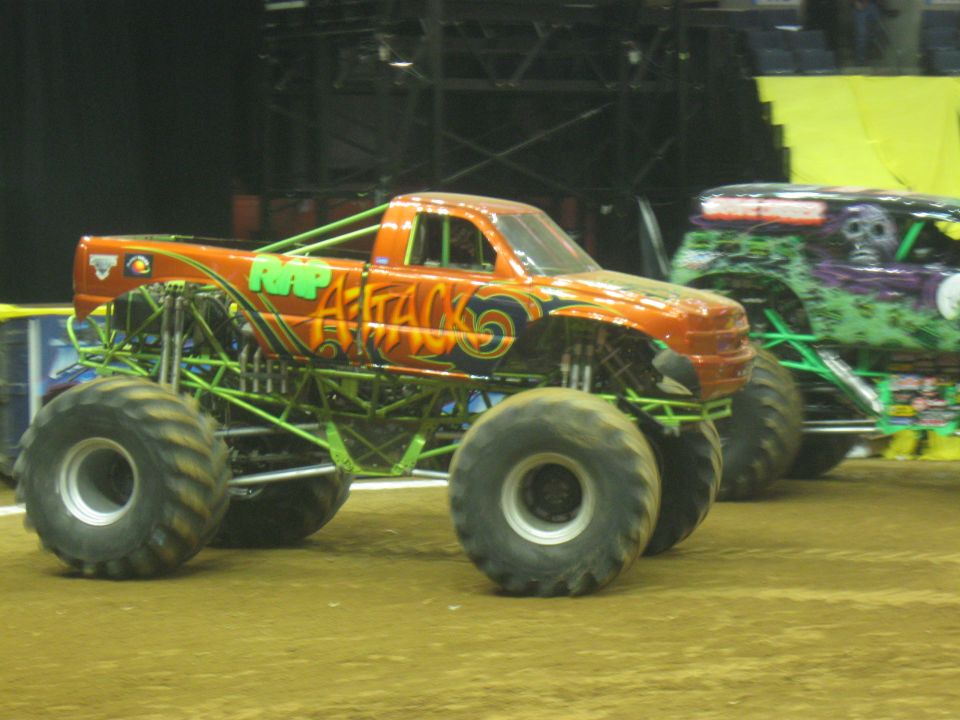 Orange monster truck with 'RAP ATTACK' on the side, next to a green monster truck with a skull graphic