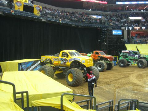 1994 Ford Bronco - Several monster trucks are lined up on a dirt arena inside a stadium, with spectators in the background.