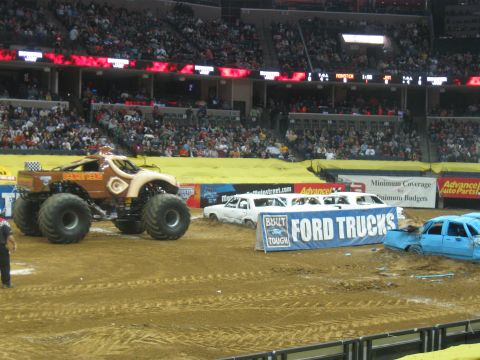 1994 Ford Bronco - A monster truck event with multiple vehicles, including a limousine and a crushed car, in a stadium arena.