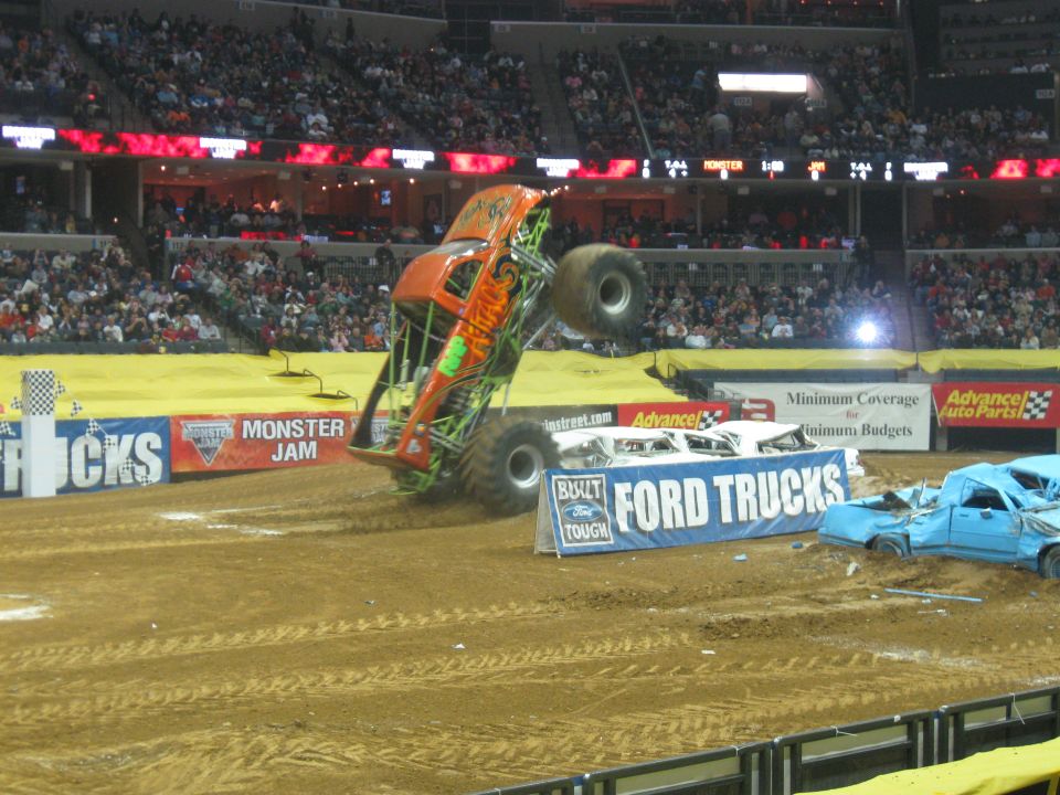 Monster truck mid-air jump over crushed cars at Monster Jam event. Ford Trucks banner visible