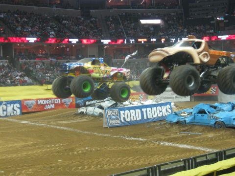 1994 Ford Bronco - Monster trucks soar over a pile of wrecked cars during a Monster Jam competition in an arena.