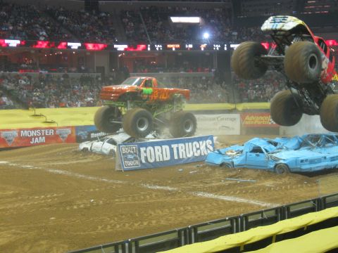 1994 Ford Bronco - Two monster trucks perform at Monster Jam, crushing cars on a dirt track under stadium lights.