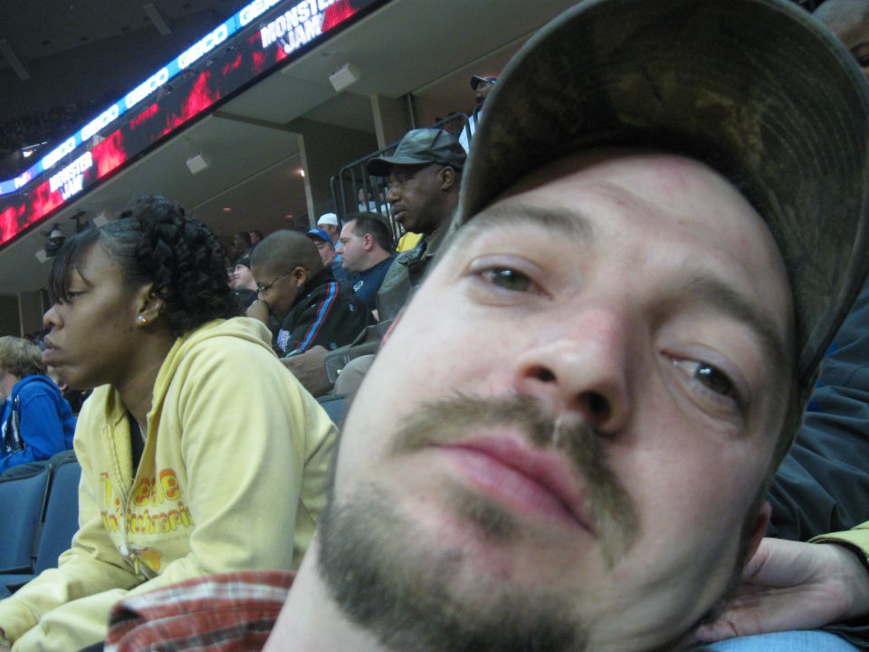 Man in foreground wearing a hat, with people in stadium seating behind him. A large screen shows 'MONSTER JAM'