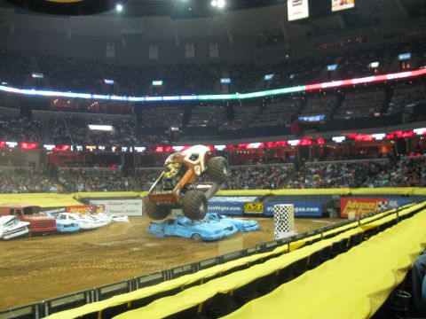 1994 Ford Bronco - A monster truck soars over a pile of wrecked cars during a competition in a packed arena.