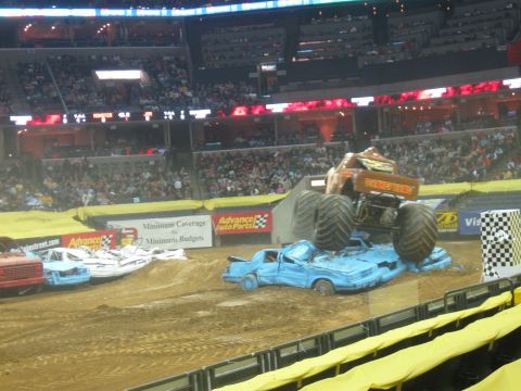 1994 Ford Bronco - A monster truck performs during an event, crushing several cars on a dirt track inside a stadium.