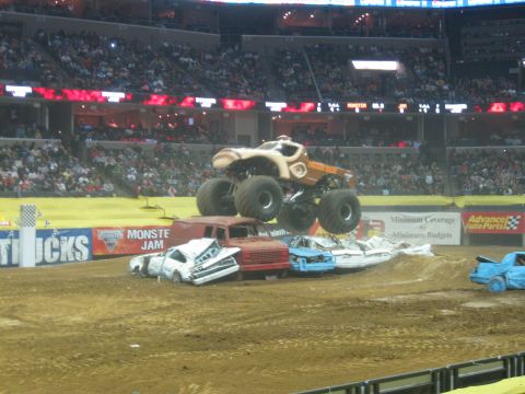 1994 Ford Bronco - A monster truck soars over a pile of mangled cars during a Monster Jam competition in a stadium.