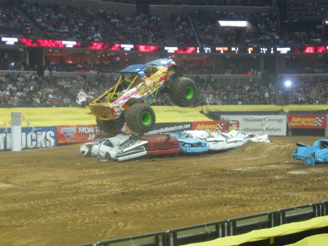 1994 Ford Bronco - A monster truck soars through the air above a pile of mangled cars during a competition.