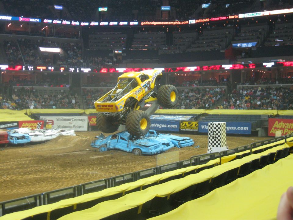 Monster truck in mid-air over crushed cars at an indoor event. Other crushed cars are visible on the ground