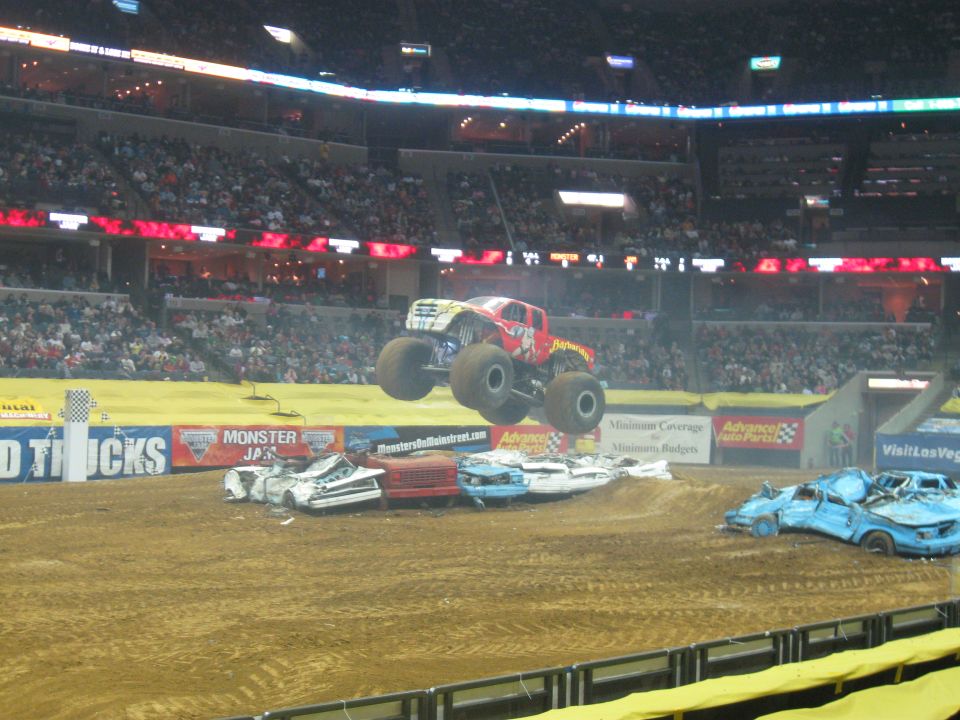 Red and white monster truck airborne over crushed cars in an arena