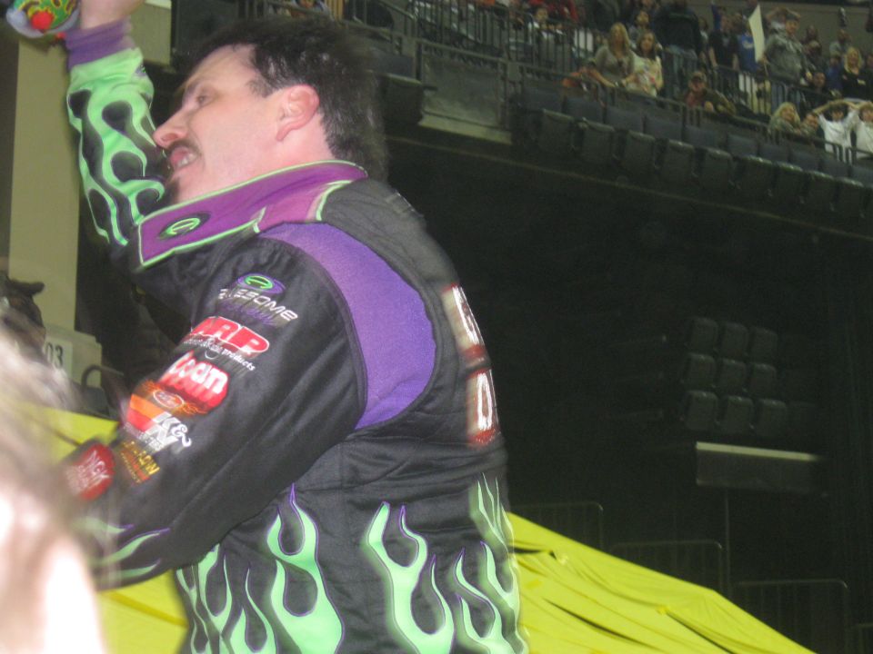 Driver in flame-patterned racing suit raises a trophy in an arena