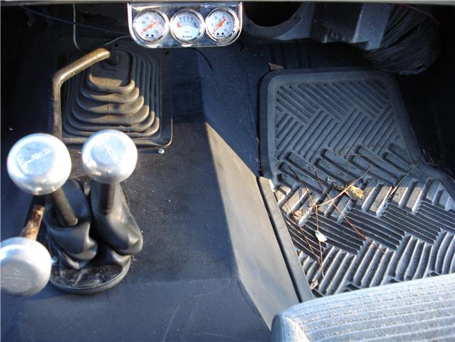 Interior shot of a vehicle's floor with two shifters, a rubber floor mat, and three gauges above