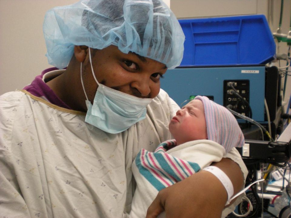 Man in surgical attire holding a newborn baby, with medical equipment in the background