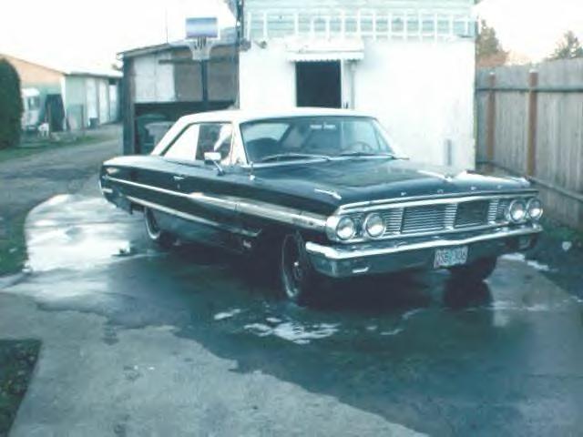 Dark vintage two-door hardtop coupe on wet driveway, 3/4 front view, white building behind