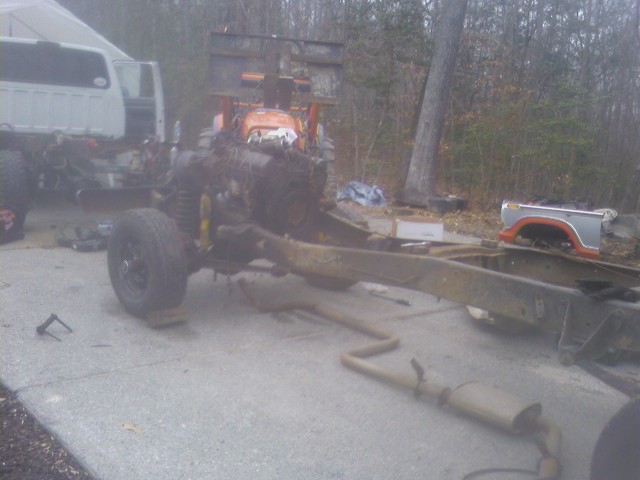 Truck frame with engine parts, a tractor behind it, and a truck bed in the background