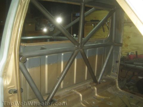 1990 Ford Bronco - Bare-metal fabricated roll cage tubing forming an X-brace fills the rear interior of a stripped vehicle cab in a workshop.