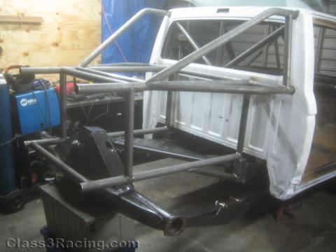 1990 Ford Bronco - bare-metal tubular space-frame chassis and roll cage are fabricated around a white truck cab on jack stands in a fab shop.