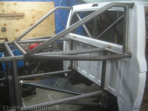 1990 Ford Bronco - Bare-metal fabricated roll cage and chassis tubing run alongside a white vehicle cab, with a welder and tools in the shop behind.