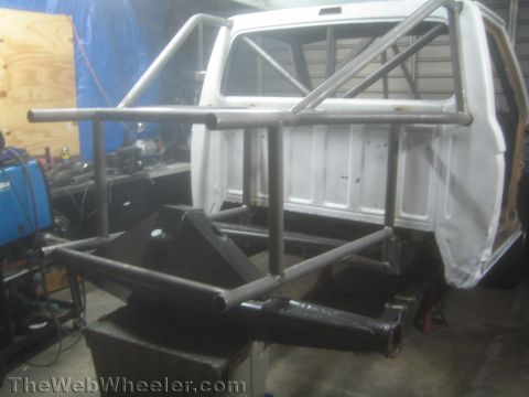 1990 Ford Bronco - bare-metal tubular chassis and roll cage with rear shock mounts is fabricated around a white truck cab on stands in a shop.