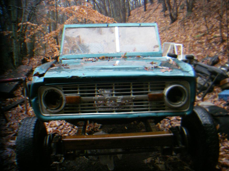 Teal 1978 Ford Bronco front grille with visible "BRONCO" lettering, missing headlights, and cracked windshield