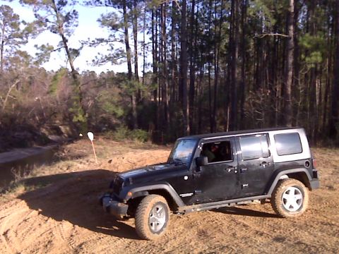 2008 Jeep Wrangler - A black four-door Jeep Wrangler is parked on a dirt path with trees in the background.