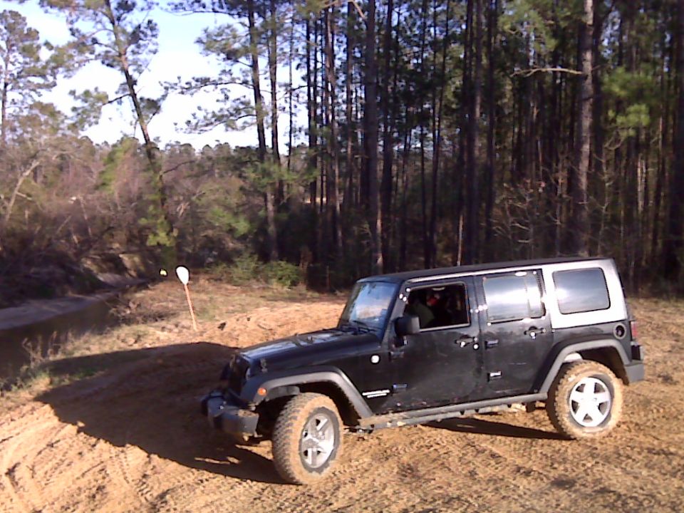 Black four-door Jeep Wrangler parked on dirt road near trees