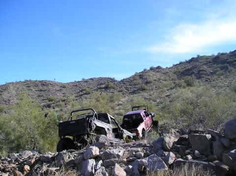 1986 Toyota Hilux - Two modified trucks sit on a rocky desert slope under a clear blue sky.