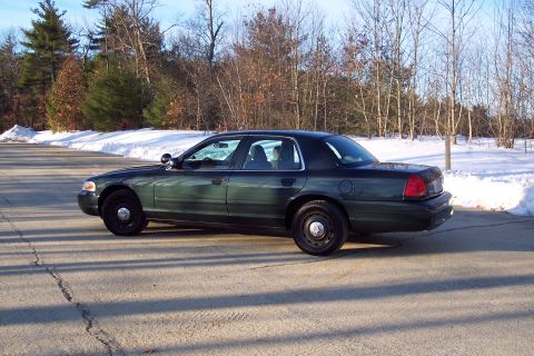 2001 Ford Crown Victoria - A dark green Ford Crown Victoria is parked on a road with snow on the ground and trees in the background.
