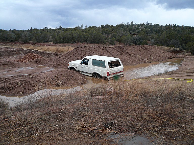 White Ford Bronco stuck in muddy water between dirt berms, seen from rear quarter