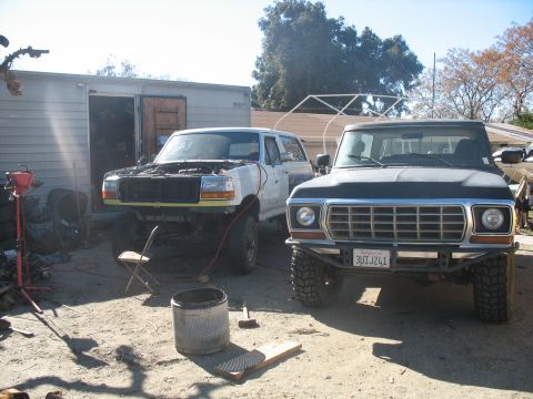 1987 Ford Bronco - Two Ford trucks, a dark F-Series and a white Bronco, are parked outside a garage.