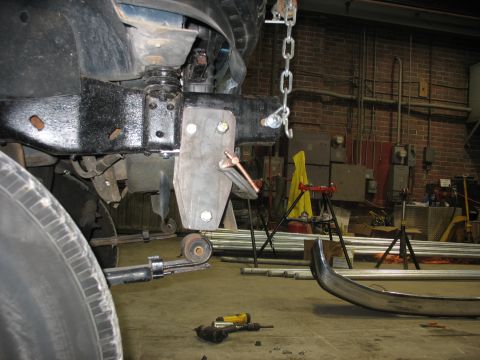 1995 Ford C-350 (Centurion Conversions) - A close-up view of a custom suspension bracket and a chain hoist being used for modification on a vehicle's frame.