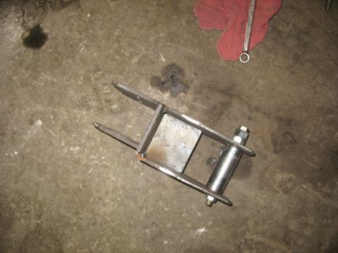 1995 Ford C-350 (Centurion Conversions) - A custom-fabricated suspension shackle assembly rests on a stained concrete surface, with a red shop rag and a wrench nearby.