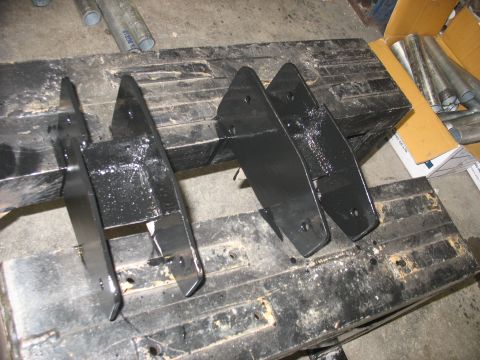 1995 Ford C-350 (Centurion Conversions) - Four black metal suspension parts are arranged on a dirty workbench, possibly for a vehicle modification.