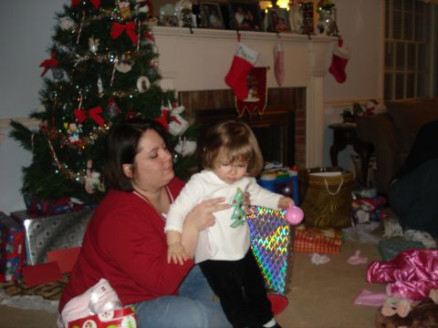 2000 Ford Crown Victoria - A woman in a red shirt is holding a young child in front of a decorated Christmas tree. The child is holding a colorful gift bag.
