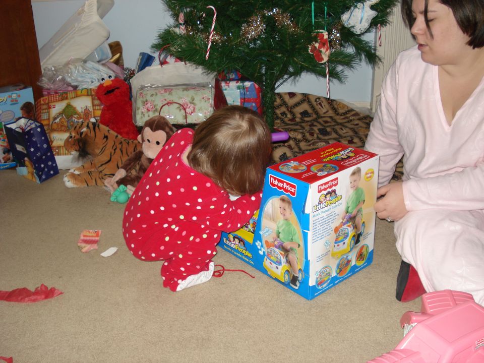 Child in red pajamas playing with toy box near Christmas tree, woman watching.