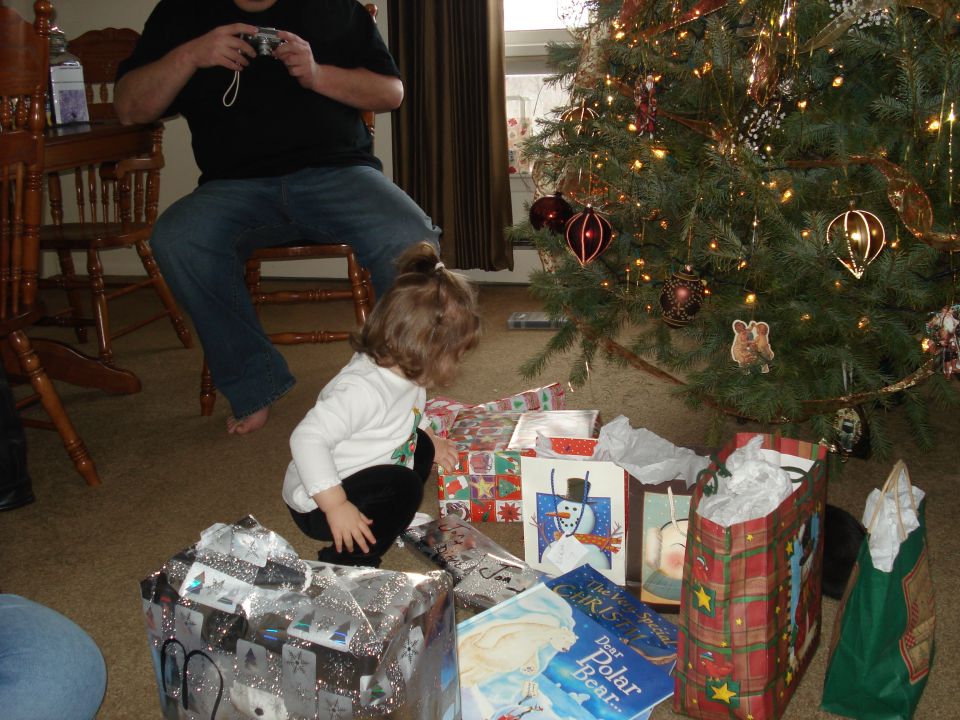A child crouches near a Christmas tree and presents, while a person holds a camera in the background.