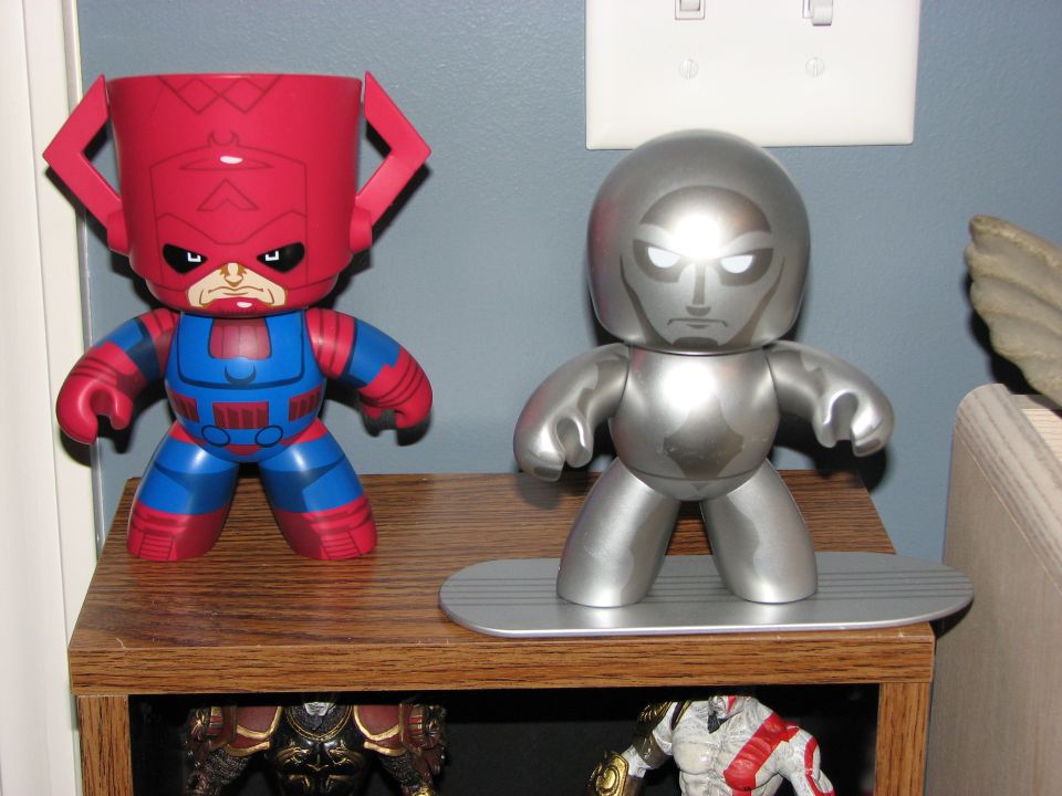 Two collectible figures, one red and blue, the other silver, stand on a wooden shelf