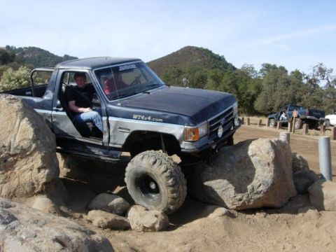 1989 Ford Bronco - Off-road truck with custom modifications navigating a rocky trail. Another vehicle is parked in the background.