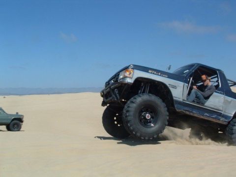 1989 Ford Bronco - A heavily modified off-road truck kicks up sand while driving on a dune, with another vehicle visible in the distance.