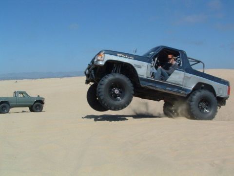 1989 Ford Bronco - Two trucks are jumping in the sand dunes under a clear blue sky.